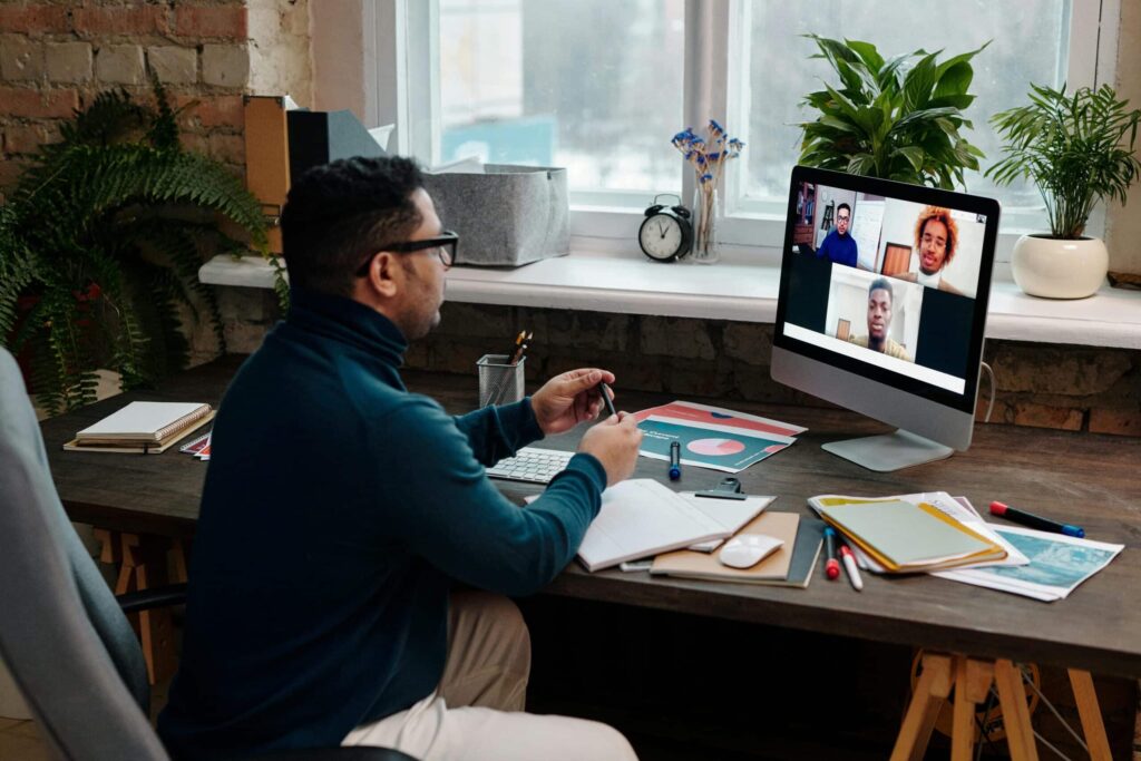A person sits at a desk with a computer, engaged in a video call. Papers and stationery are scattered on the workspace, surrounded by plants.