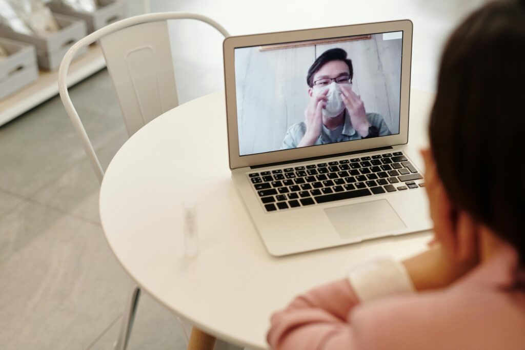 A person sitting at a white table, attentively engaging in a video call on a laptop. Bright indoor environment with minimalist decor.