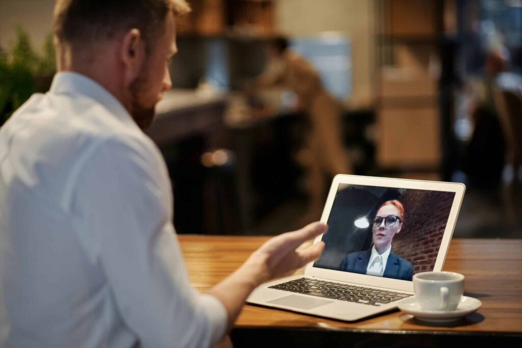 A man in a white shirt gestures while video chatting on a laptop at a cozy café, with a coffee cup on the wooden table.