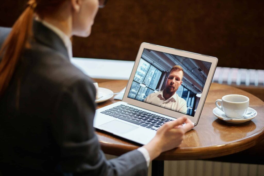 A woman in a blazer sits at a café table, engaging in a video call on her laptop, with coffee next to her.