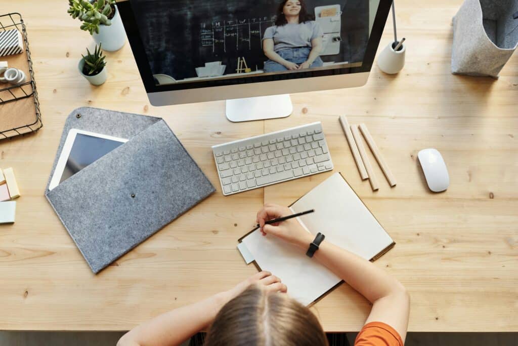 A workspace with a person writing in a notebook, a computer monitor displaying a video, and various office supplies on a wooden desk.