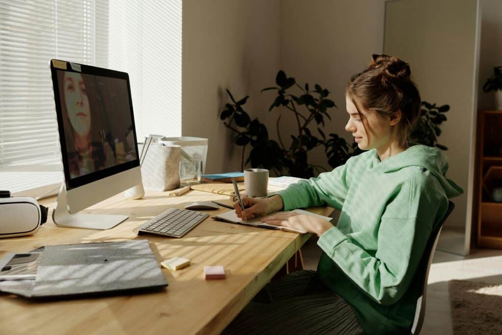 A person in a green hoodie writes in a notebook at a wooden desk, with a computer and plants in a bright, sunlit room.