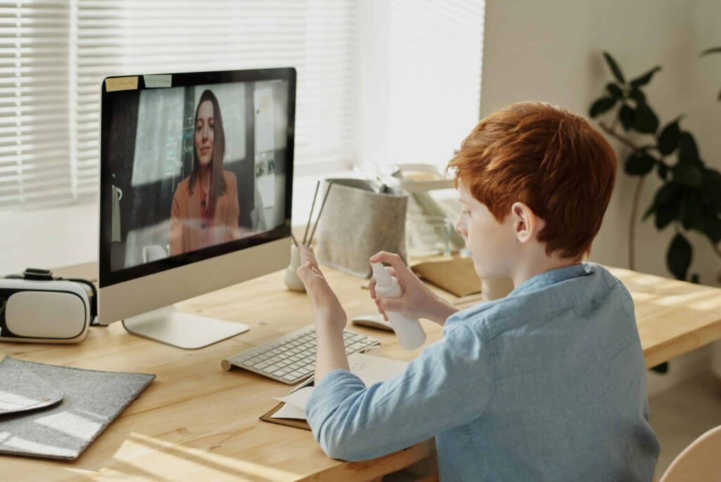 A child with red hair sits at a wooden desk, holding a spray bottle while engaging in a video call on a computer.