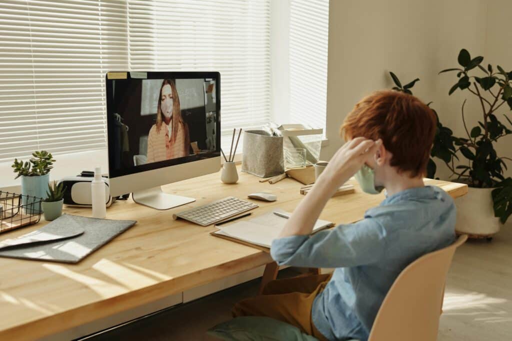A person with red hair sits at a wooden desk, attentively watching a video call on a computer in a bright, minimalistic office.