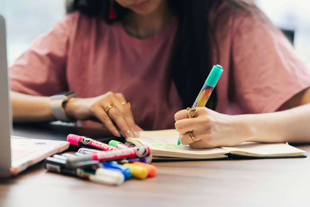 A person wearing a pink shirt writes in a notebook with colorful markers scattered on a wooden table. A laptop is visible nearby.
