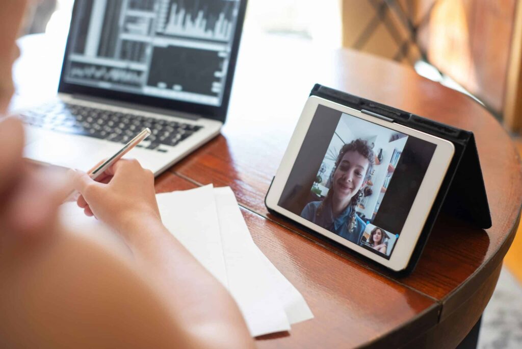 A person taking notes while engaged in a video call on a tablet, with a laptop displaying data analysis in the background.