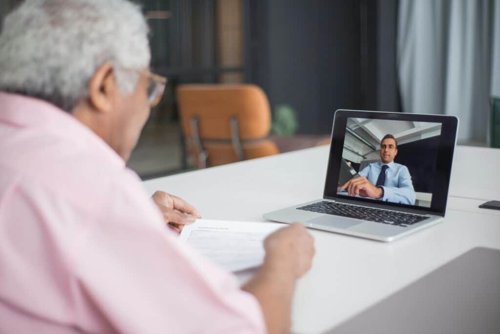 An elderly person is engaged in a video call on a laptop, holding documents and focused on the screen.