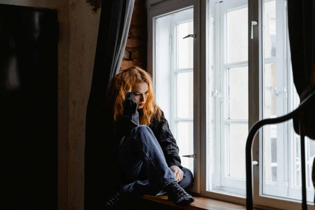 A person with wavy red hair sits curled up on a windowsill, gazing out the window, framed by dark curtains and exposed brick.