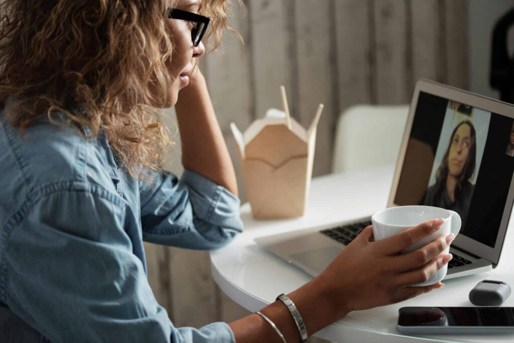 A person with curly hair sits at a table, holding a white mug while video chatting on a laptop, with takeout nearby.