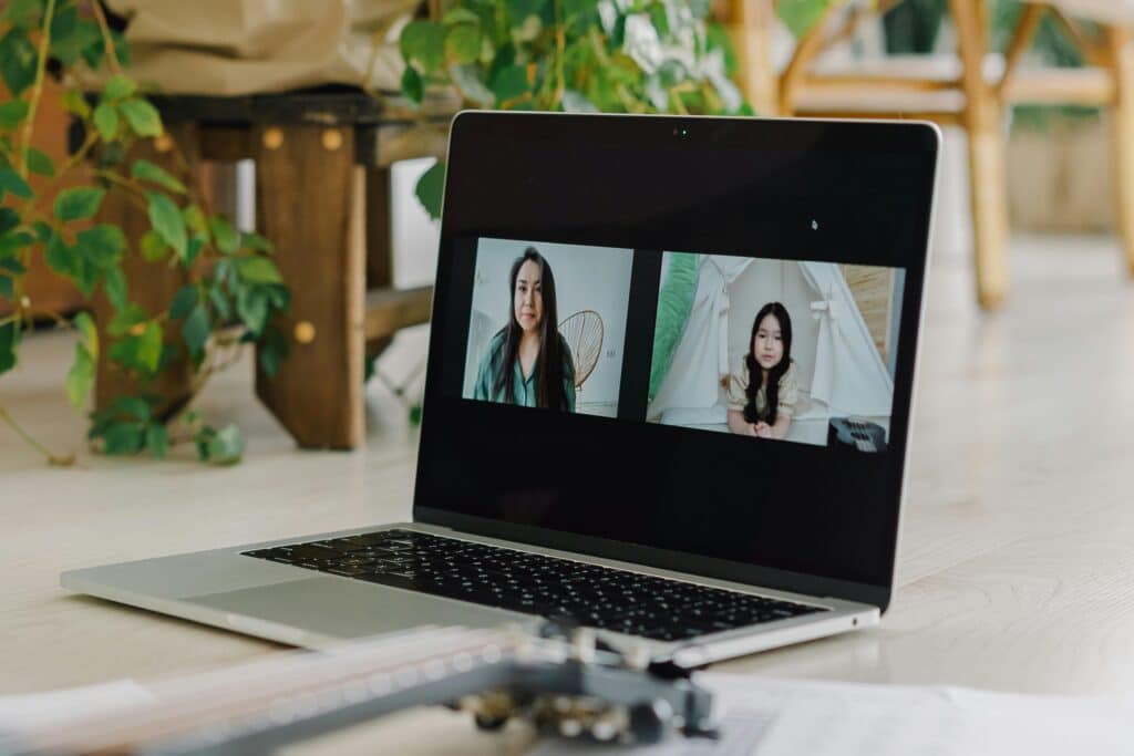 A laptop on a wooden floor displays two video call participants. A guitar and sheets of music are visible in the foreground.