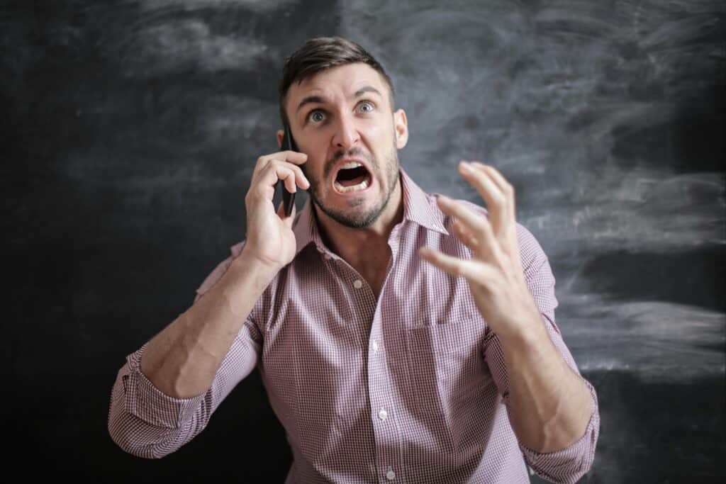 A man in a checkered shirt gestures animatedly while talking on the phone against a dark, chalkboard-like background.