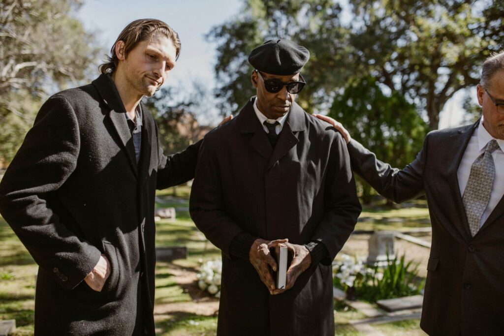 Three individuals in formal attire stand together outdoors in a cemetery, with one holding a book, offering support to one another.