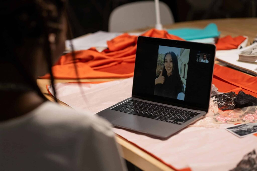 A person sitting at a table with a laptop, engaged in a video call, surrounded by colorful fabric and sewing materials.
