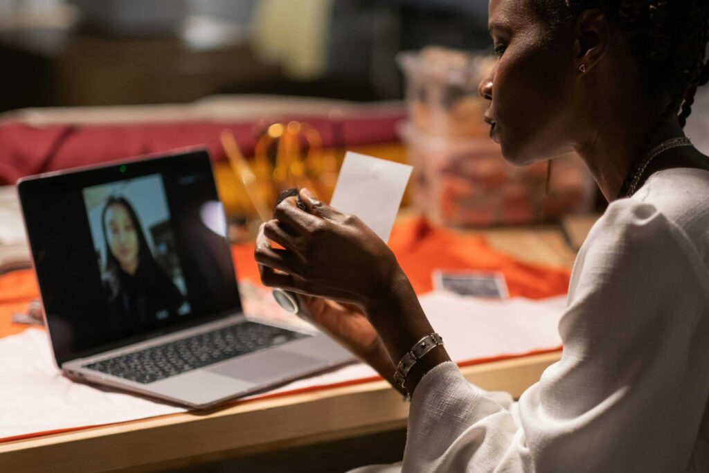 A person holds a card while engaging in a video call on a laptop, surrounded by colorful fabric and various items on a table.