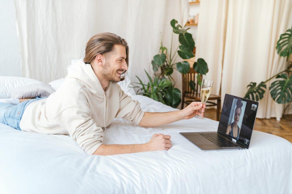 A person in a cream hoodie lies on a bed, holding a champagne glass while video chatting on a laptop, surrounded by indoor plants.