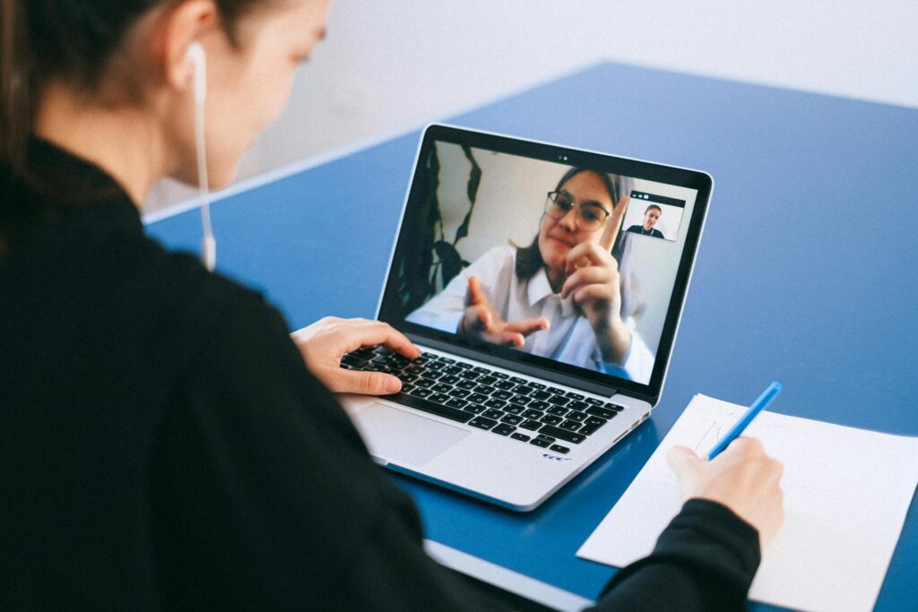 A person participates in a video call on a laptop, writing notes on paper beside them, with a blue tabletop in the background.