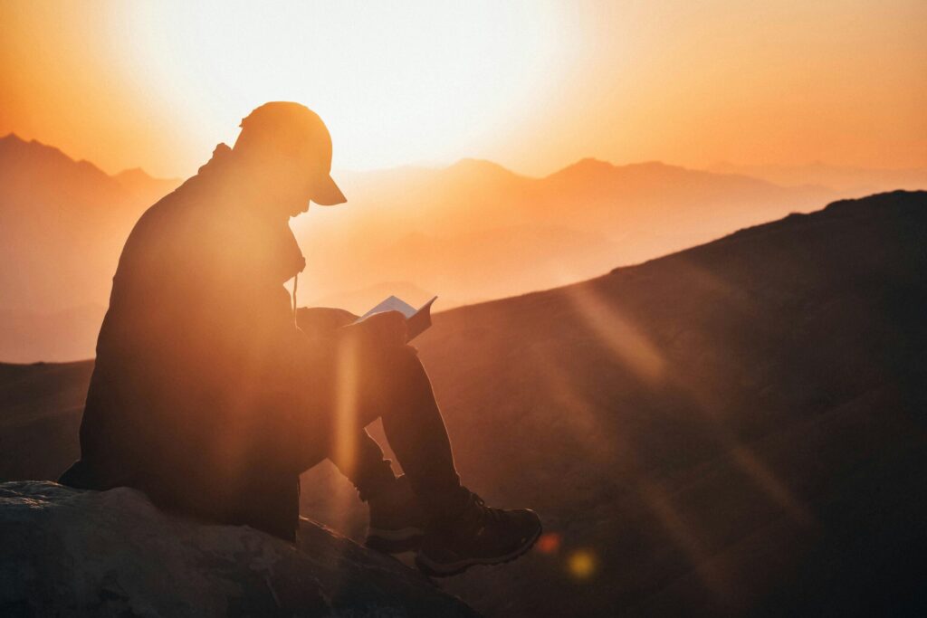 Silhouette of a person reading a book at sunset, surrounded by mountains, with warm sunlight creating a serene atmosphere.