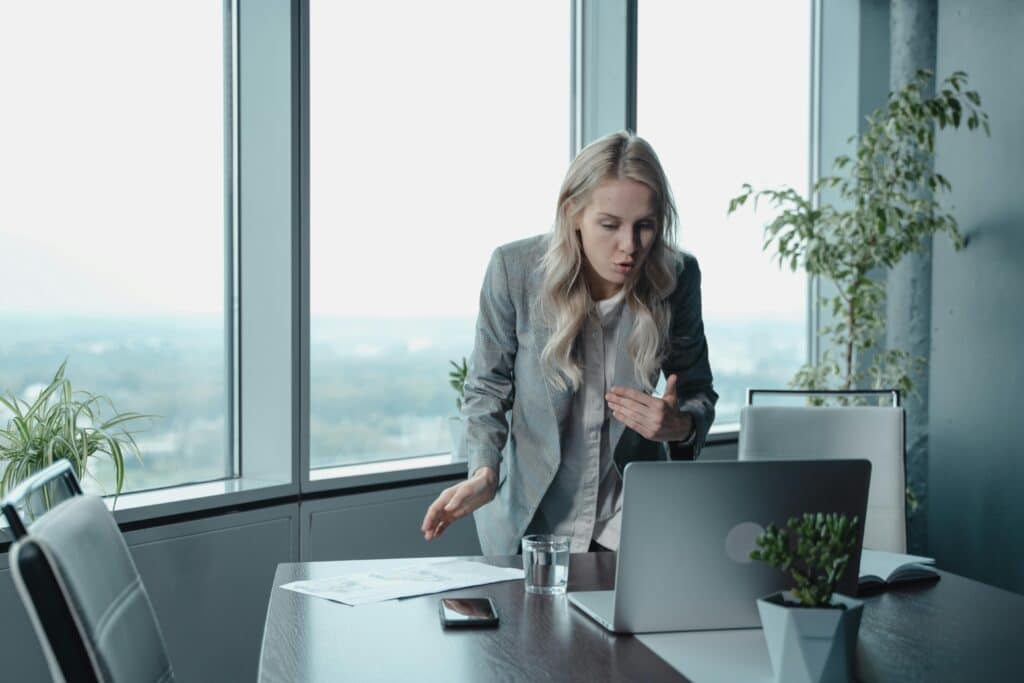 A businesswoman in a neat blazer stands by a conference table, gesturing toward documents and a laptop with a city view behind her.