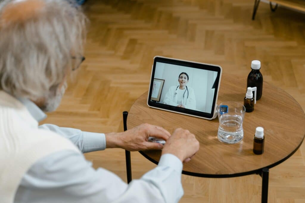 An elderly person is seated at a table, interacting with a doctor on a tablet, surrounded by bottles of medicine and a glass of water.