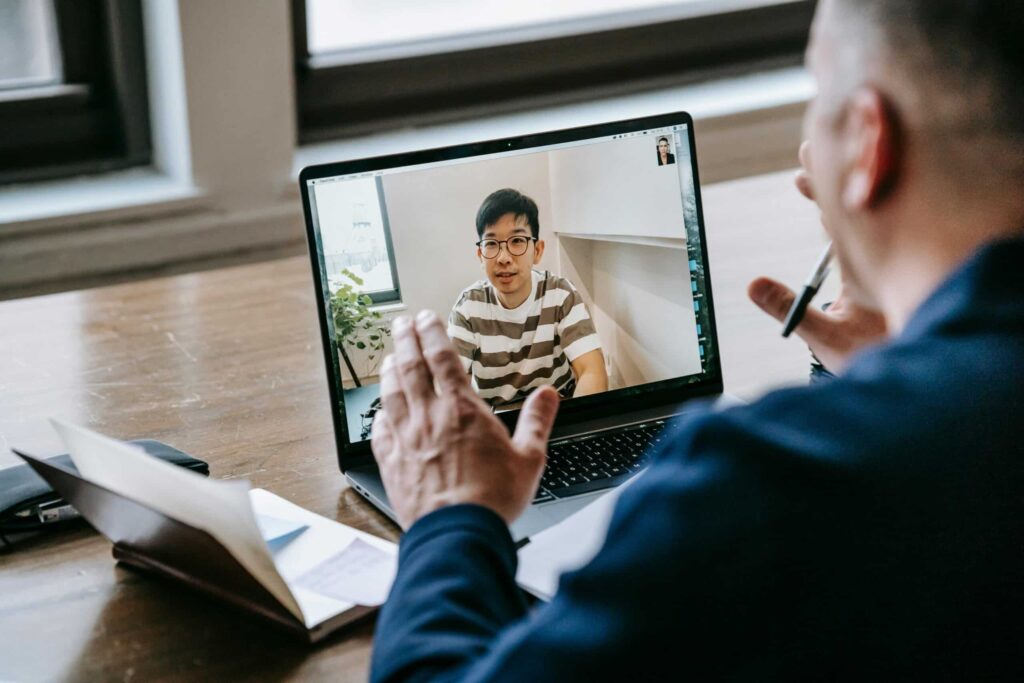 A person participates in a video call on a laptop, using hand gestures while seated at a wooden table with notes and a phone.