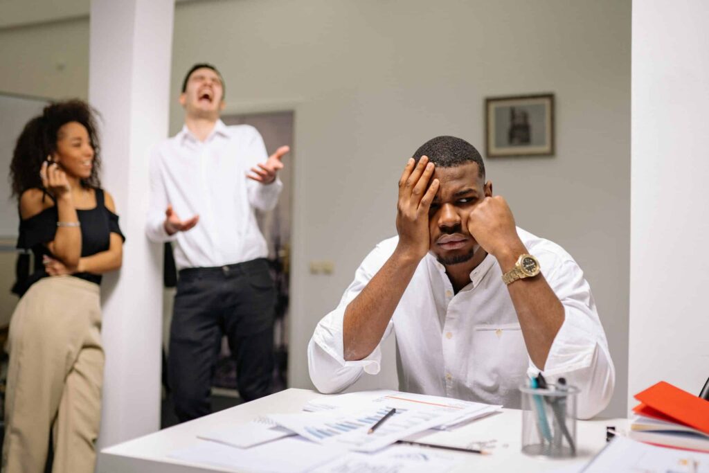 A man sits at a desk, appearing stressed, while a woman stands nearby with her arms crossed. Another person gestures expressively.
