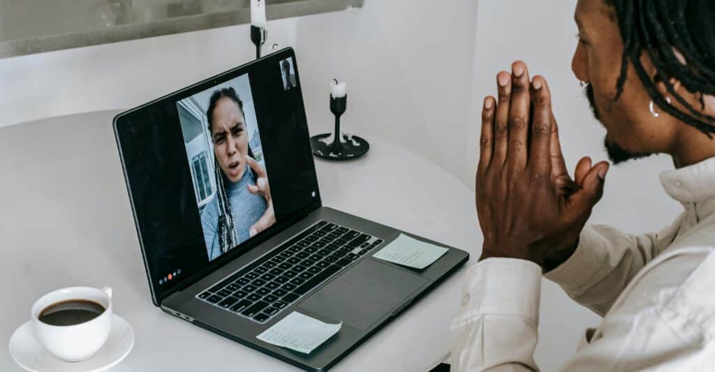 A person with braided hair sits at a table with a laptop showing a video call, a cup of coffee, and sticky notes nearby.