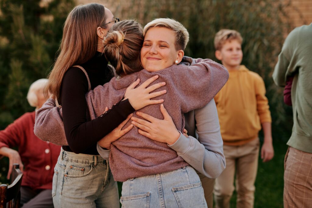 Two women share a warm embrace in a joyful outdoor setting, surrounded by people engaging in conversation in the background.