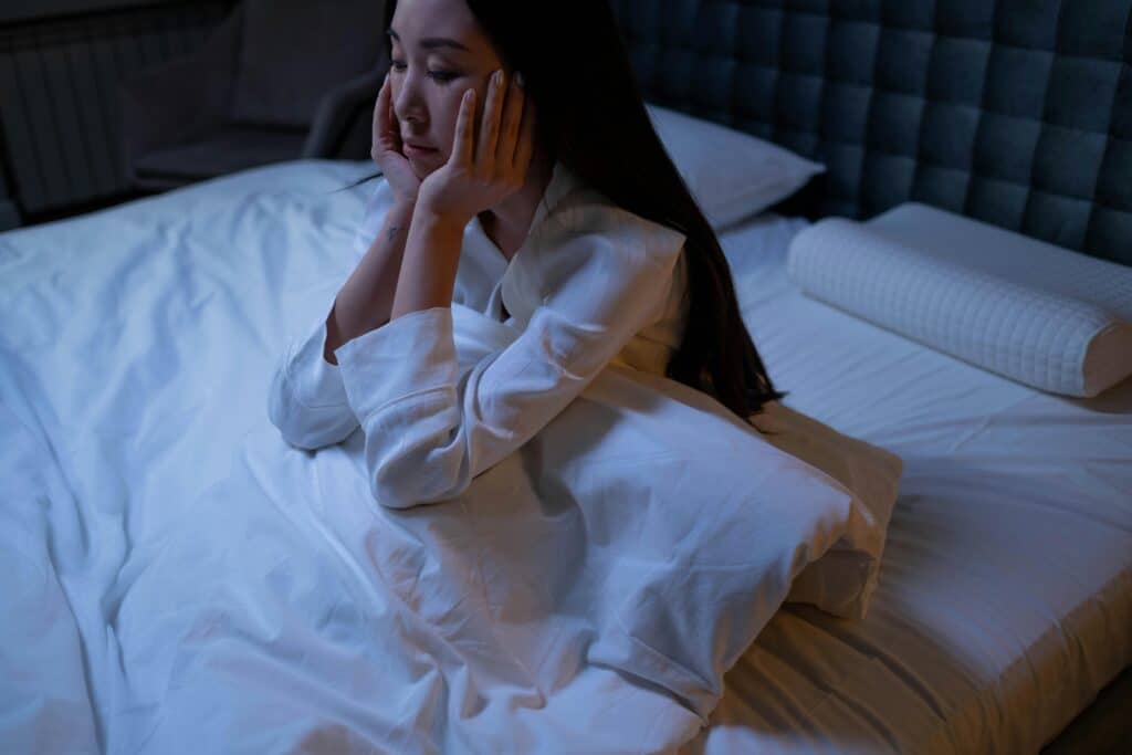 A person sits on a bed, wrapped in white sheets, with their hands covering their face, in a dimly lit room.