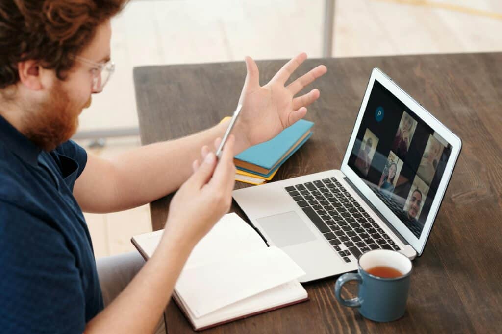 A man with curly hair gestures while speaking in a video call on a laptop. An open notebook, colored folders, and a cup of tea are on the table.