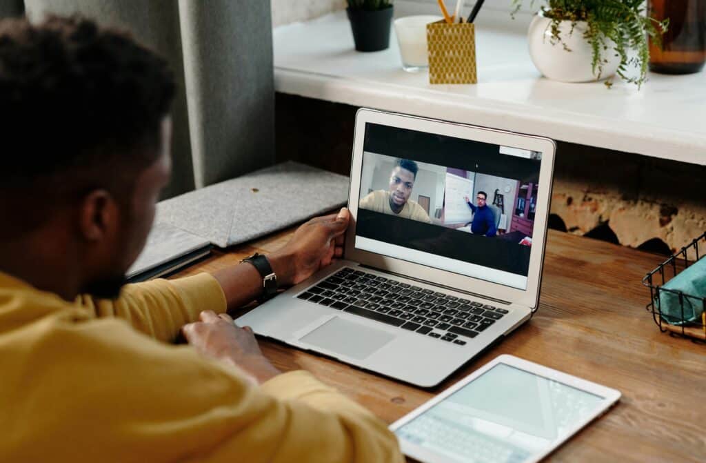 A person in a yellow shirt sits at a desk, participating in a video call on a laptop with a tablet beside them.