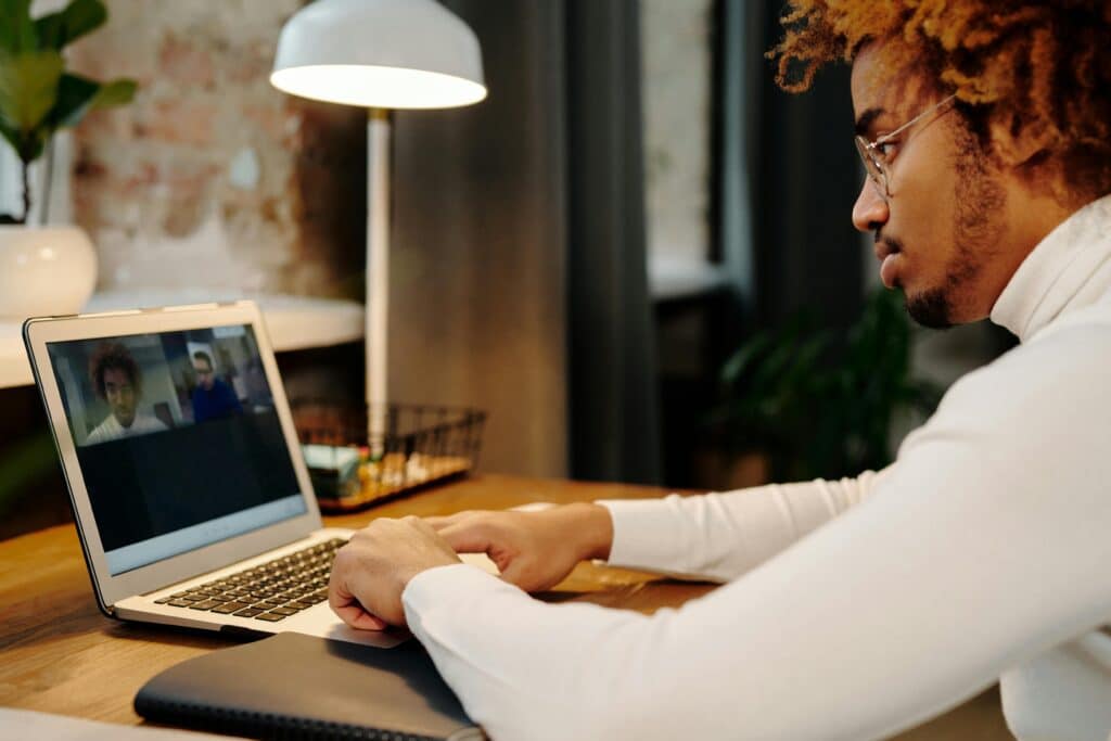 A person in a white turtleneck is engaged in a video call on a laptop at a wooden desk, with a stylish lamp and plant in the background.