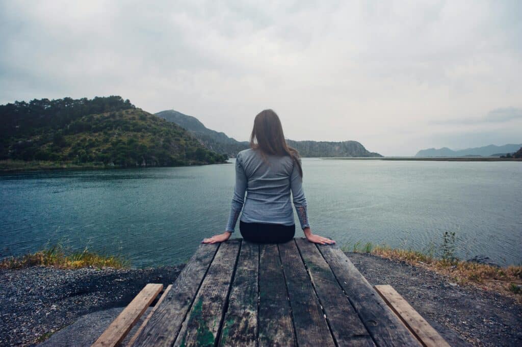 A woman sits on a weathered dock, gazing serenely over a calm lake surrounded by hills under a cloudy sky.