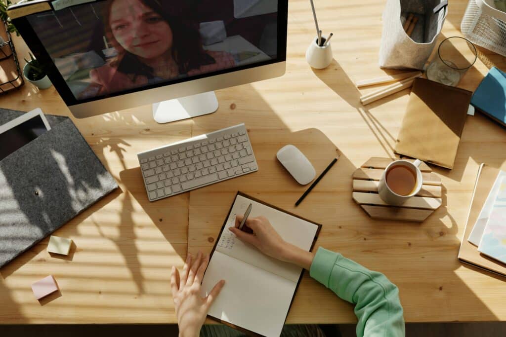 A workspace with a computer, notebook, and coffee cup; sunlight casts shadows on a wooden desk adorned with stationery and plants.