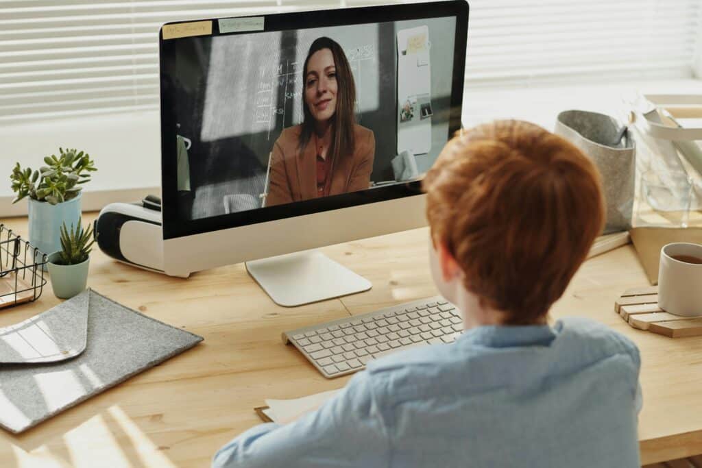 A child with red hair sits at a desk, engaged in an online class via a desktop computer surrounded by plants and stationery.