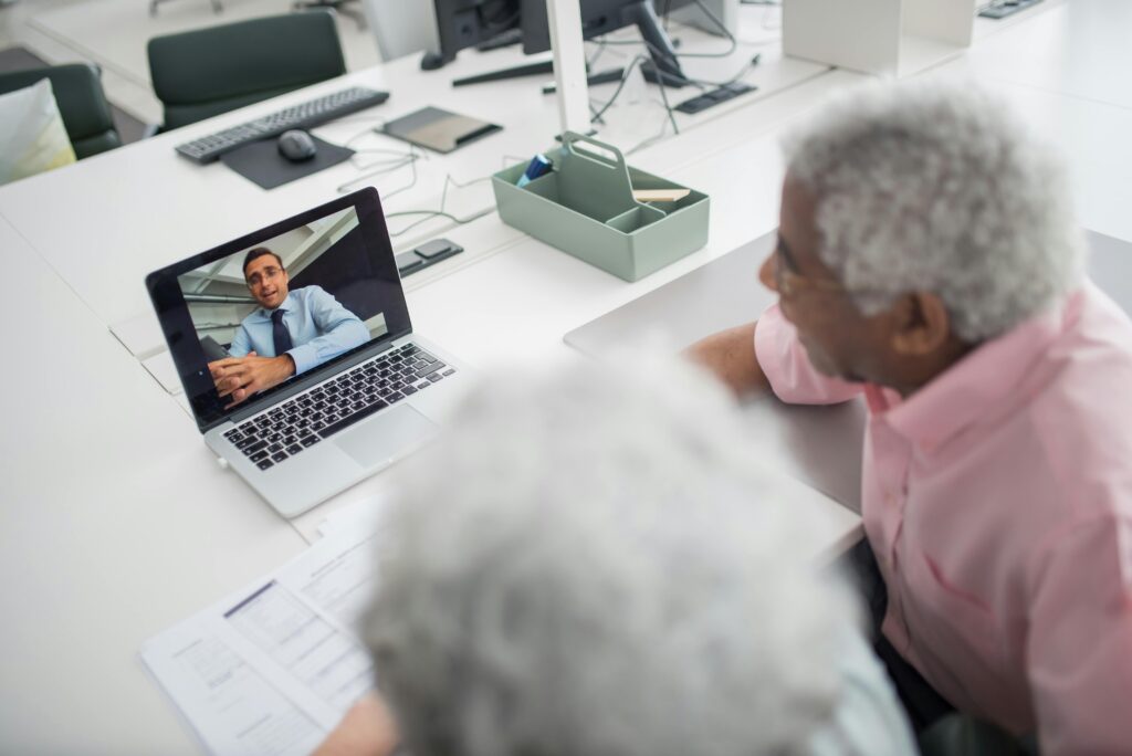 An elderly couple engages in a video call on a laptop at a modern workspace, accompanied by documents and office supplies.