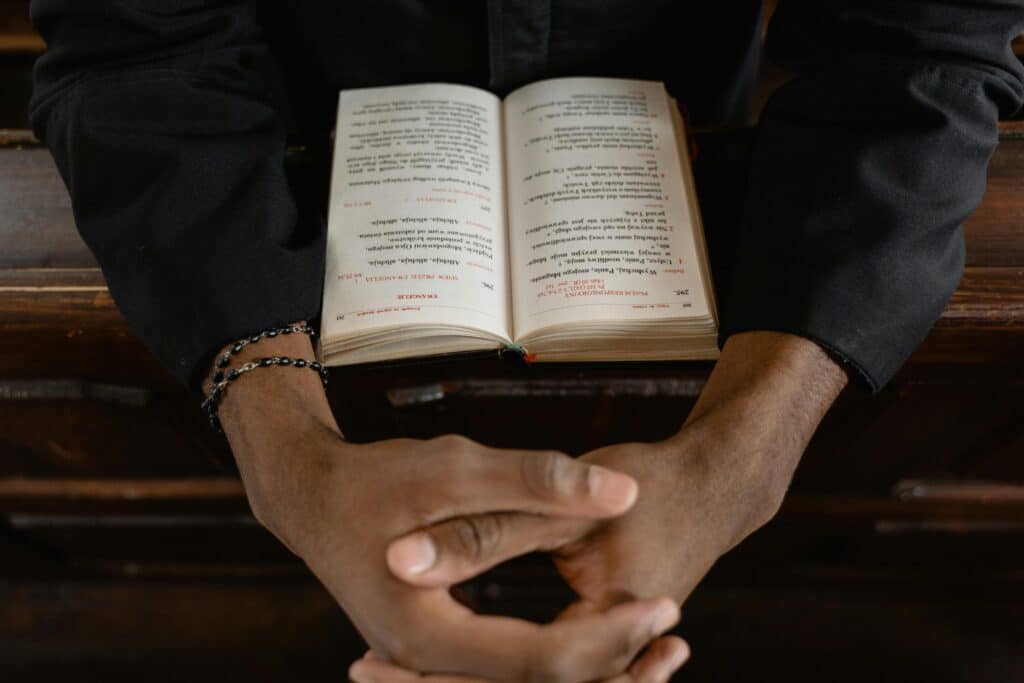A pair of hands clasped together in prayer above an open book, resting on a wooden surface, conveying contemplation and spirituality.