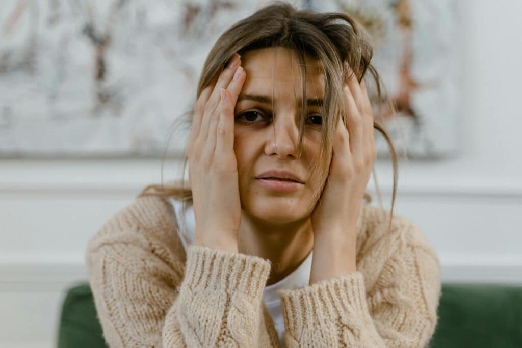 A person sits on a green sofa, holding their head in their hands, wearing a cozy beige sweater, against a neutral background.