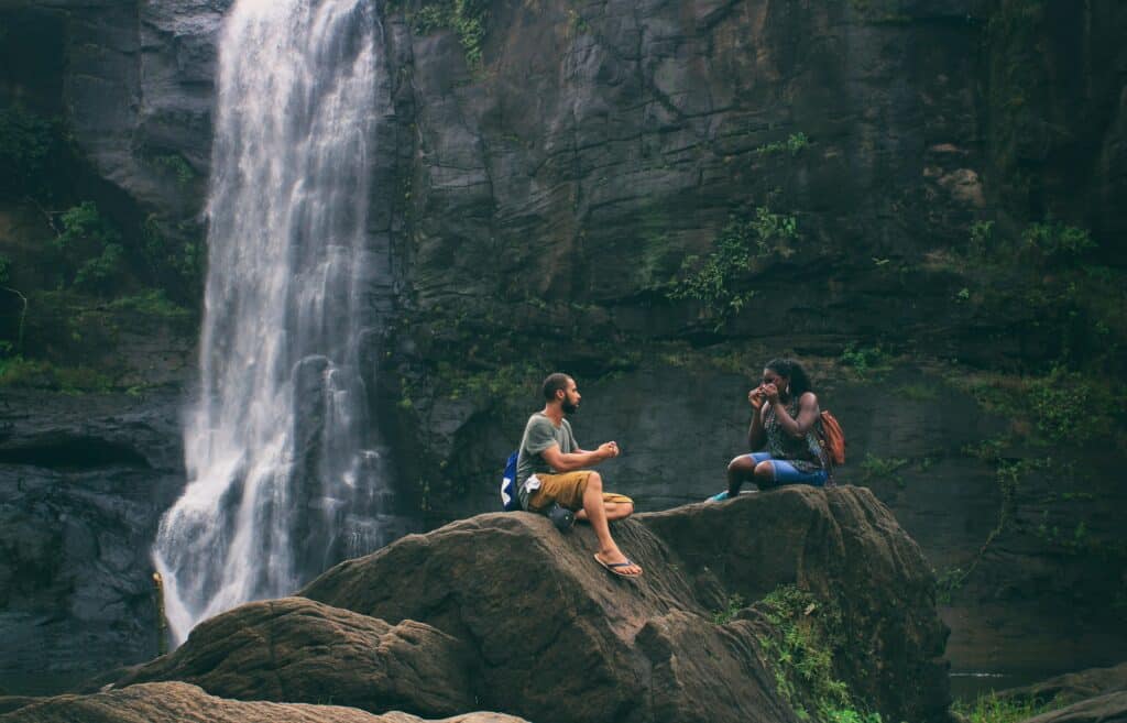 Two individuals sit on large rocks in front of a cascading waterfall, surrounded by lush greenery and a serene natural landscape.