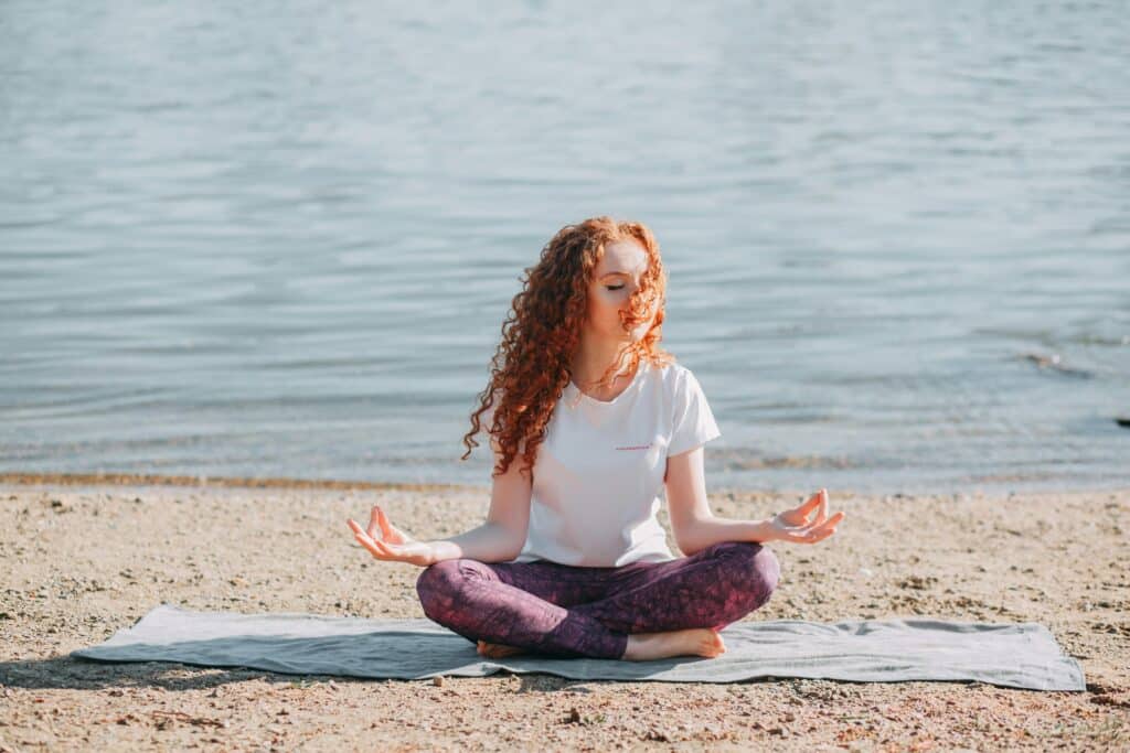 A person with curly hair sits cross-legged on a mat by the water, practicing meditation in a serene outdoor setting.