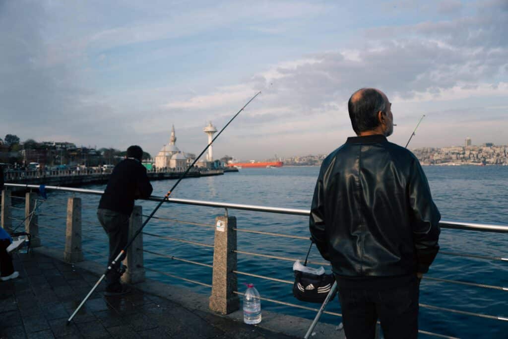 Two fishermen stand on a pier, one casting a line into the water while the other gazes toward the horizon, with cityscapes in view.