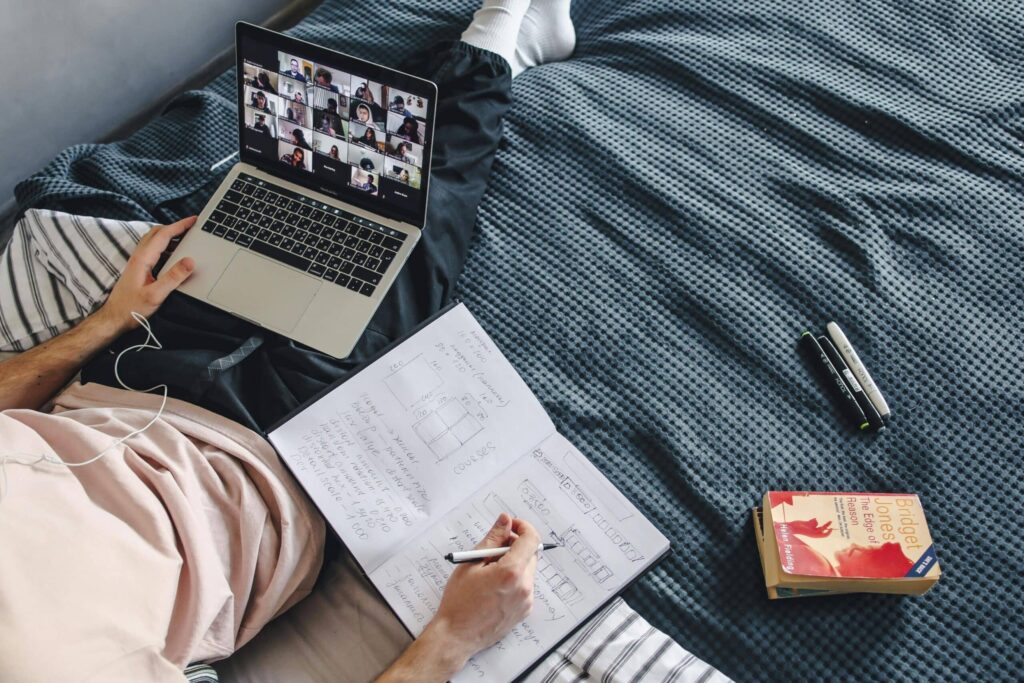 A person sits on a bed with a laptop displaying a video call, taking notes in a notebook beside a book and pens.