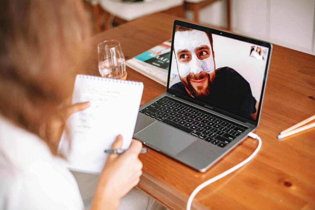 A person is engaged in a video call on a laptop while taking notes in a notebook, with a glass of water nearby.