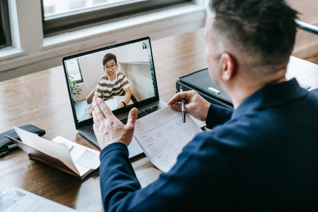 A person participates in a virtual meeting, gesturing, while studying notes on a laptop at a wooden table with documents nearby.