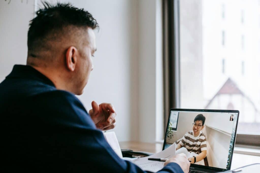 A person in a suit gestures during a video call, with a laptop displaying a seated individual in a casual outfit.