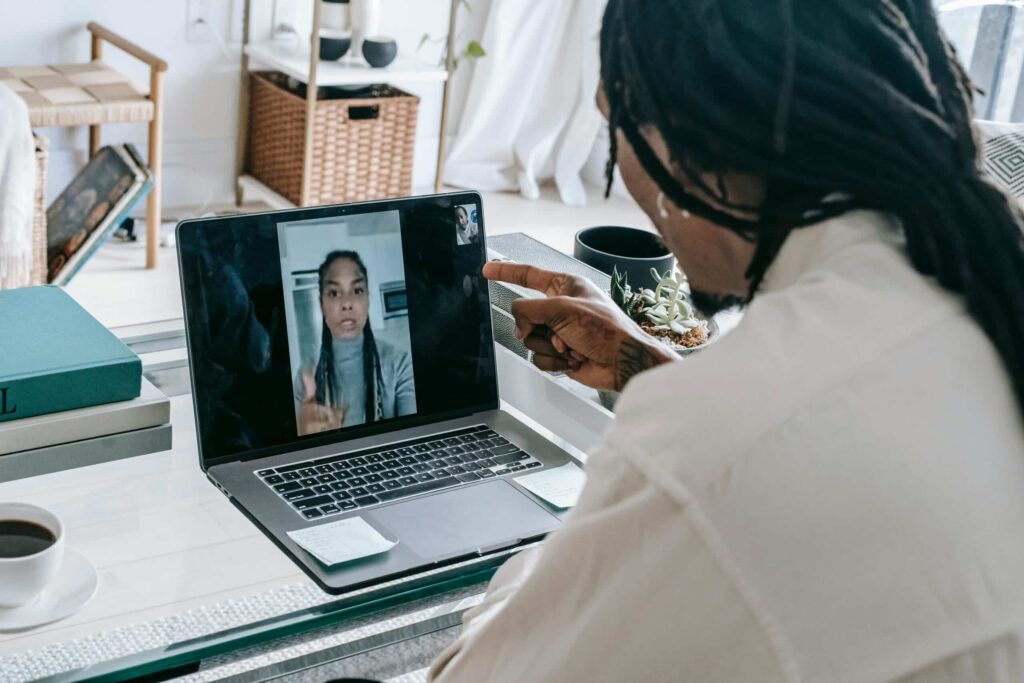 A person with dreadlocks gestures while on a video call, seated at a glass table with coffee and succulent plants around.