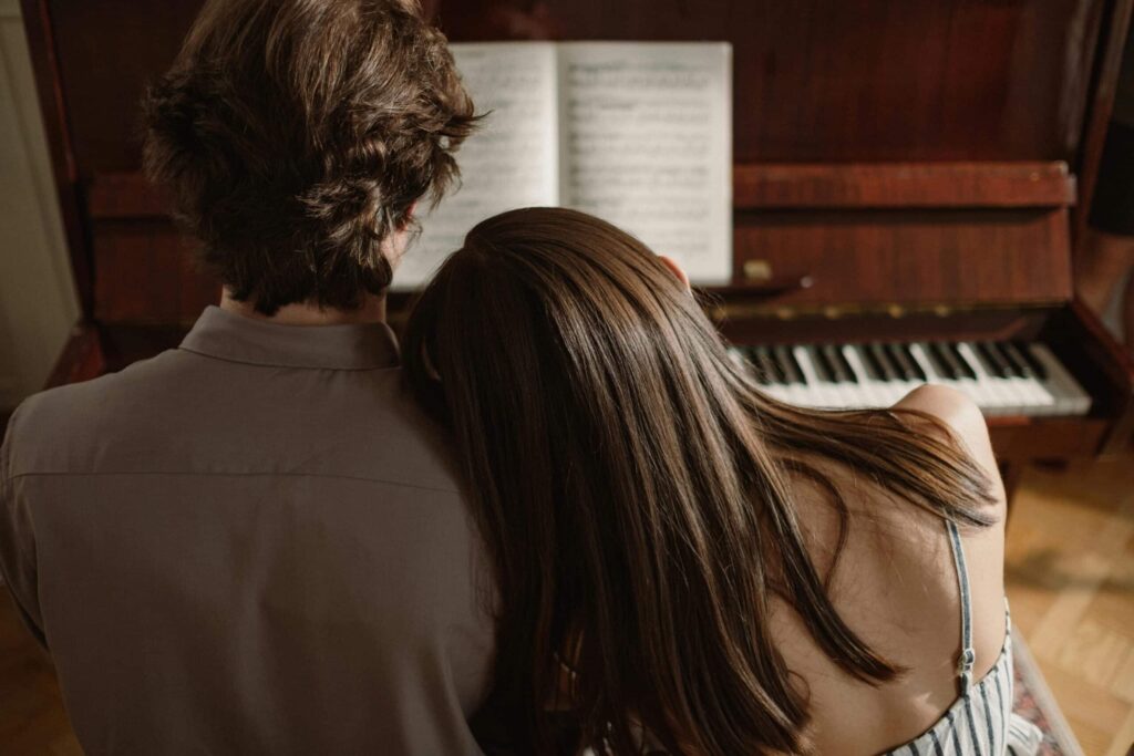 A couple sits closely at a piano, with the woman resting her head on the man's shoulder, sharing a moment of intimacy.
