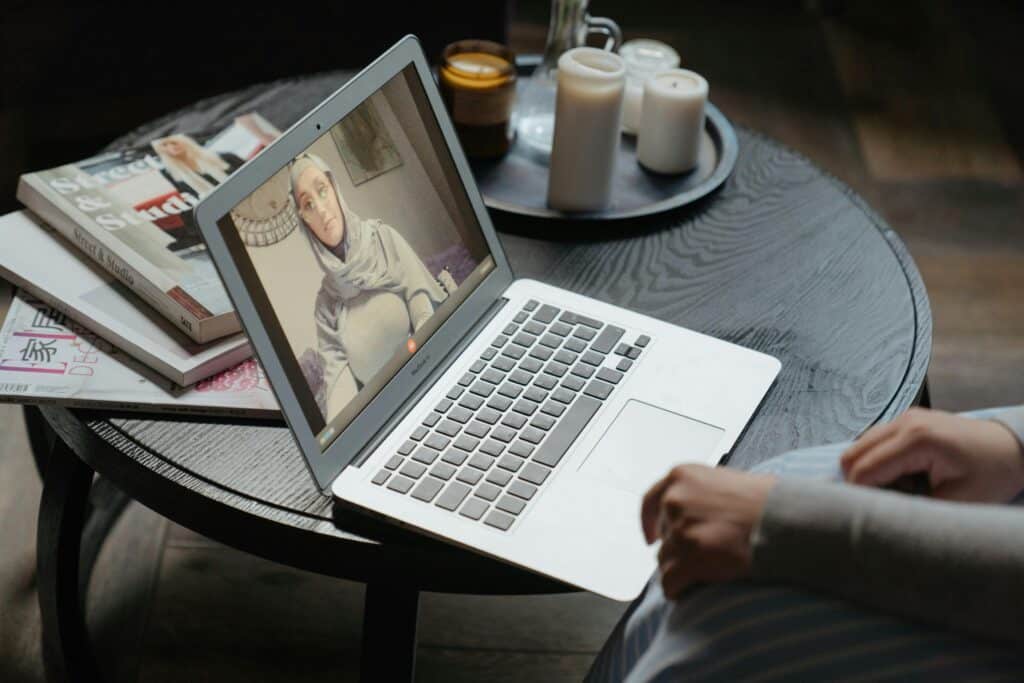 A person sits at a wooden table, engaged in a video call on a laptop surrounded by magazines and candles.