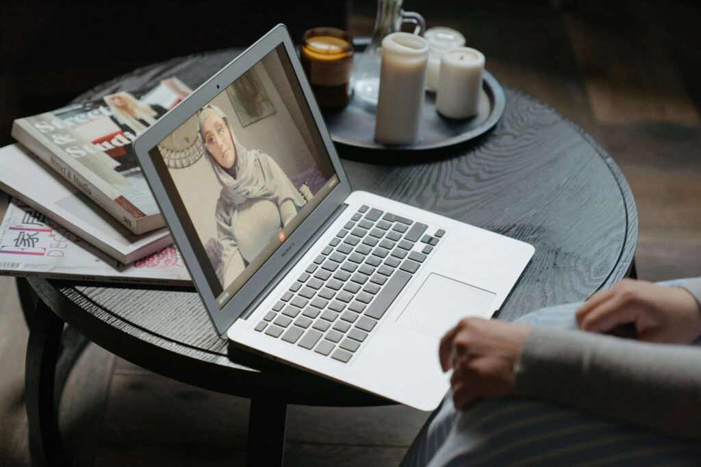 A person sits at a wooden table, engaged in a video call on a laptop surrounded by magazines and candles.