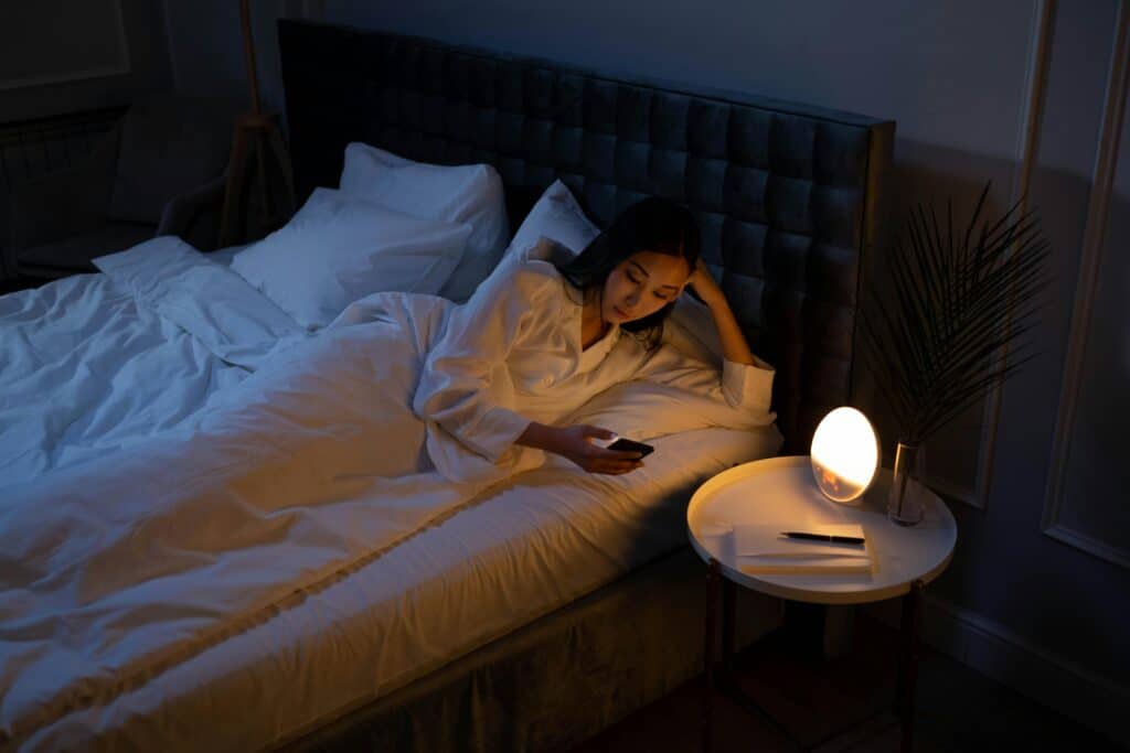 A person in a white robe lies in bed, browsing a smartphone, with soft lighting from a lamp on a nearby side table.