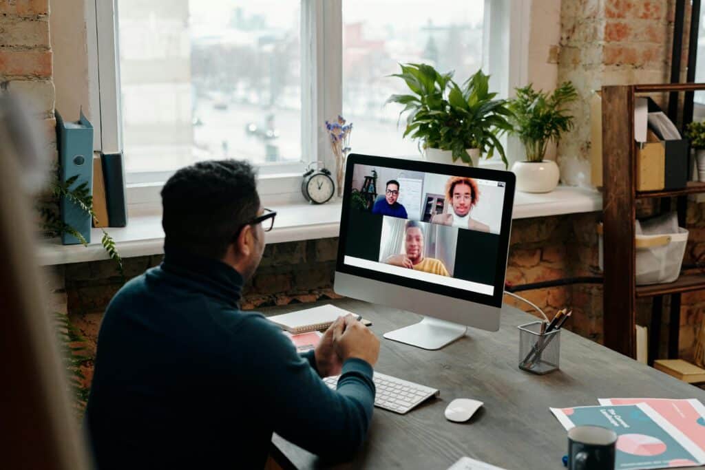 A man sits at a desk, participating in a video call on his computer, with plants and office supplies around him, and a city view outside.